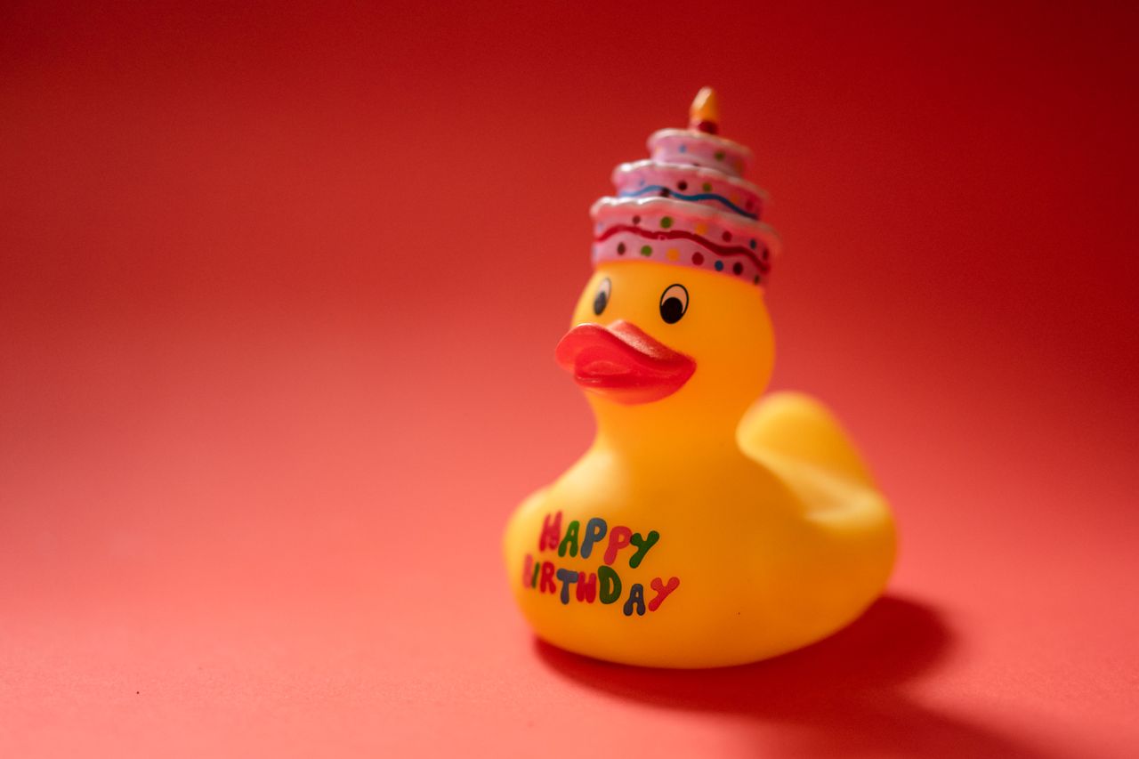 A yellow rubber duck wearing a birthday cake hat, with HAPPY BIRTHDAY printed on its side, against a red background