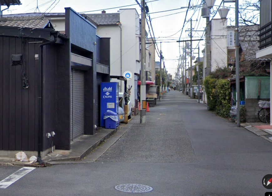 A vending machine at the road edge beside a dark wooden building in Hino