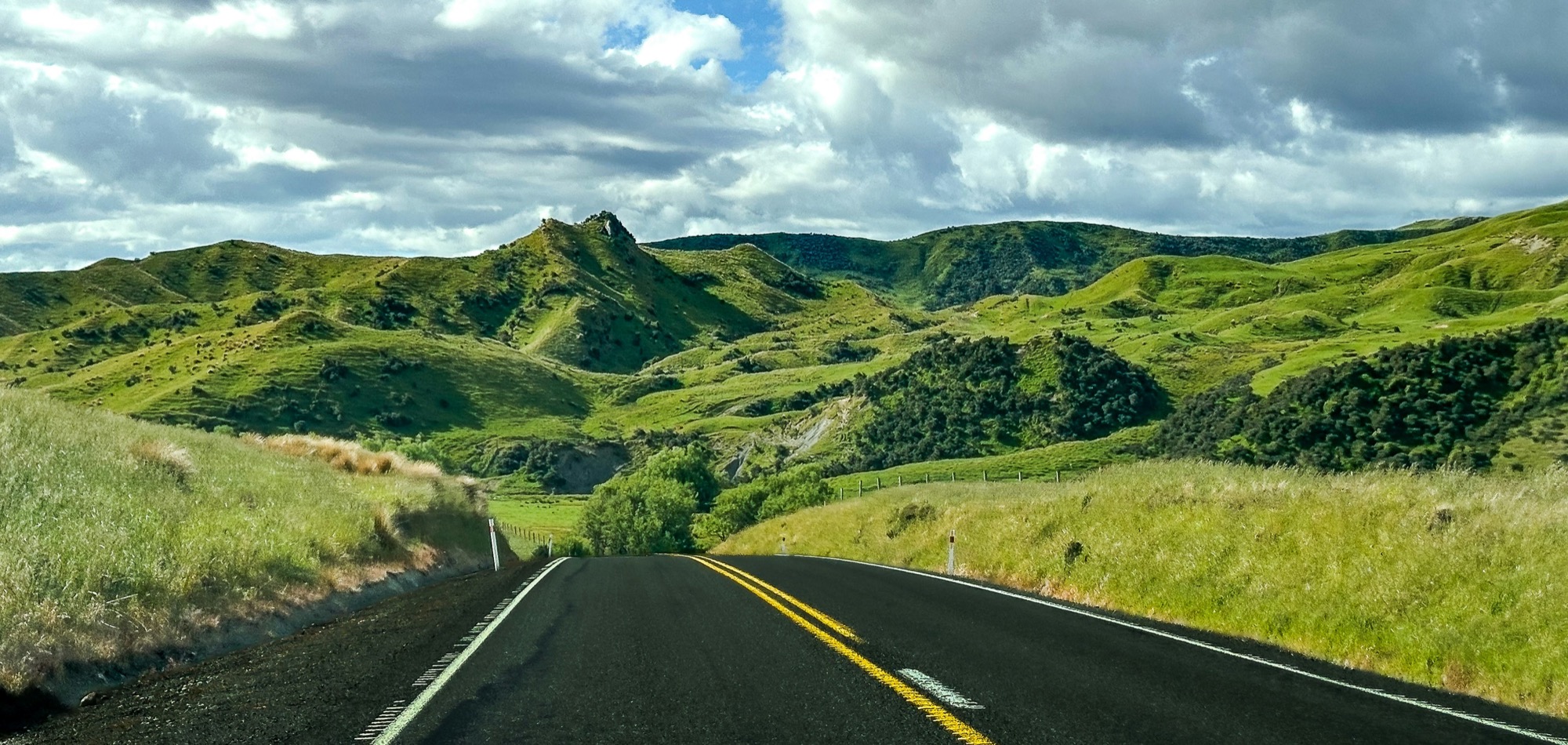 The South Island: made for a slow tour on four wheels or two An empty road curving through bright green rolling hills under a big cloudy sky - open-road touring in New Zealand