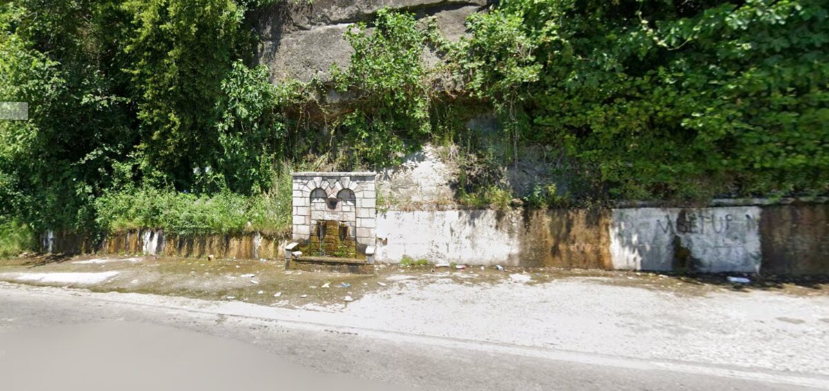 A stone arched roadside fountain with water flowing from two spouts, set into a wall beneath a rock face