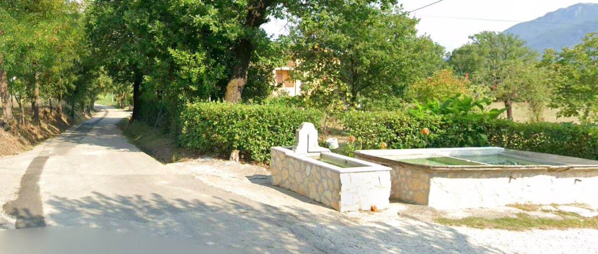 A white stone trough fountain with a spout beside a quiet rural lane in Italy