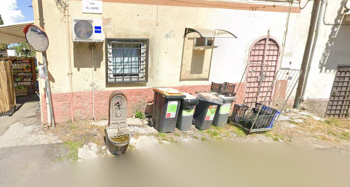 A classic cast-iron Italian drinking fountain on a street corner between wheelie bins, on Via del Lavatoio